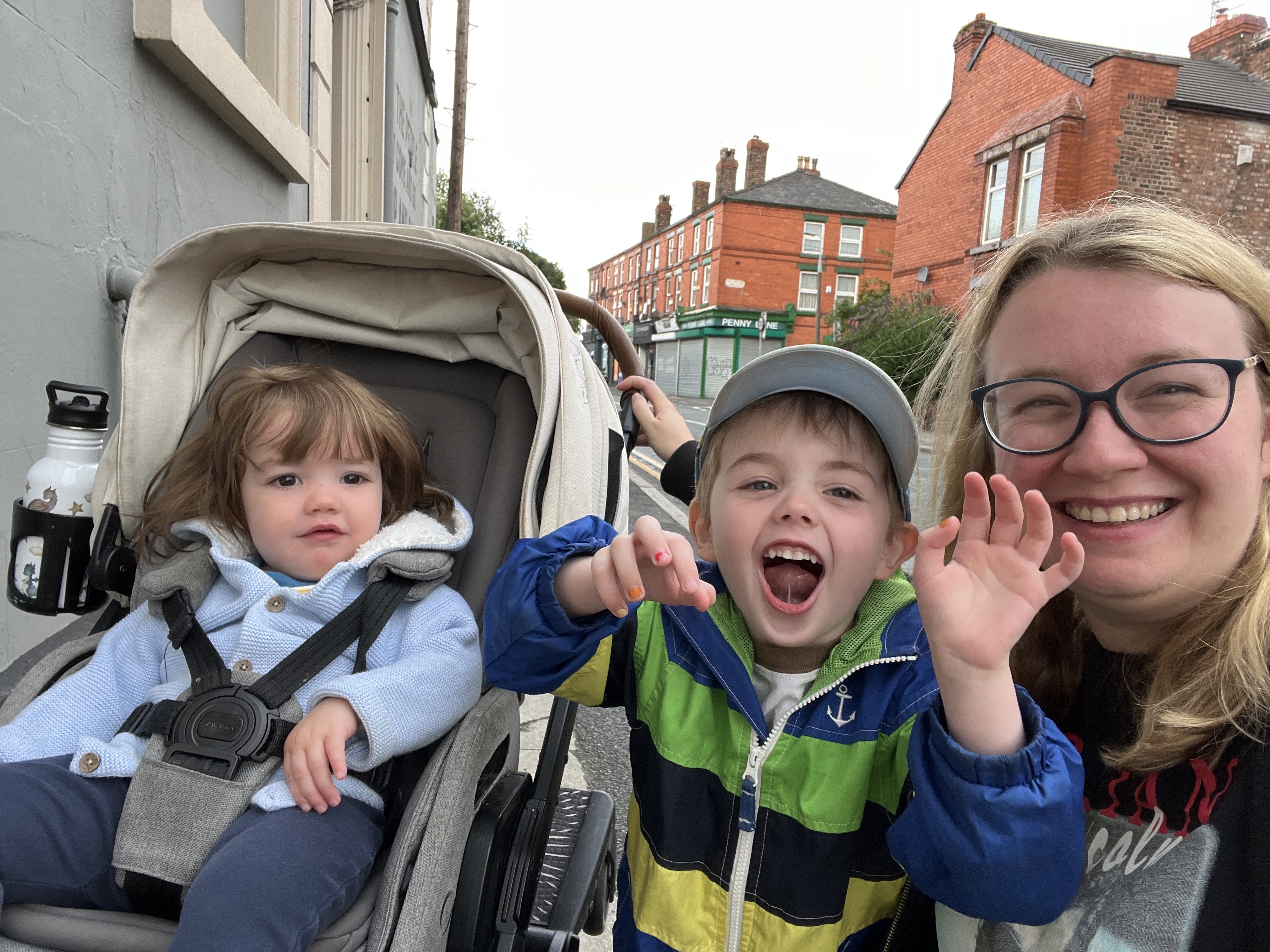 A woman with two young children; one is in a stroller, the other is standing next to her.