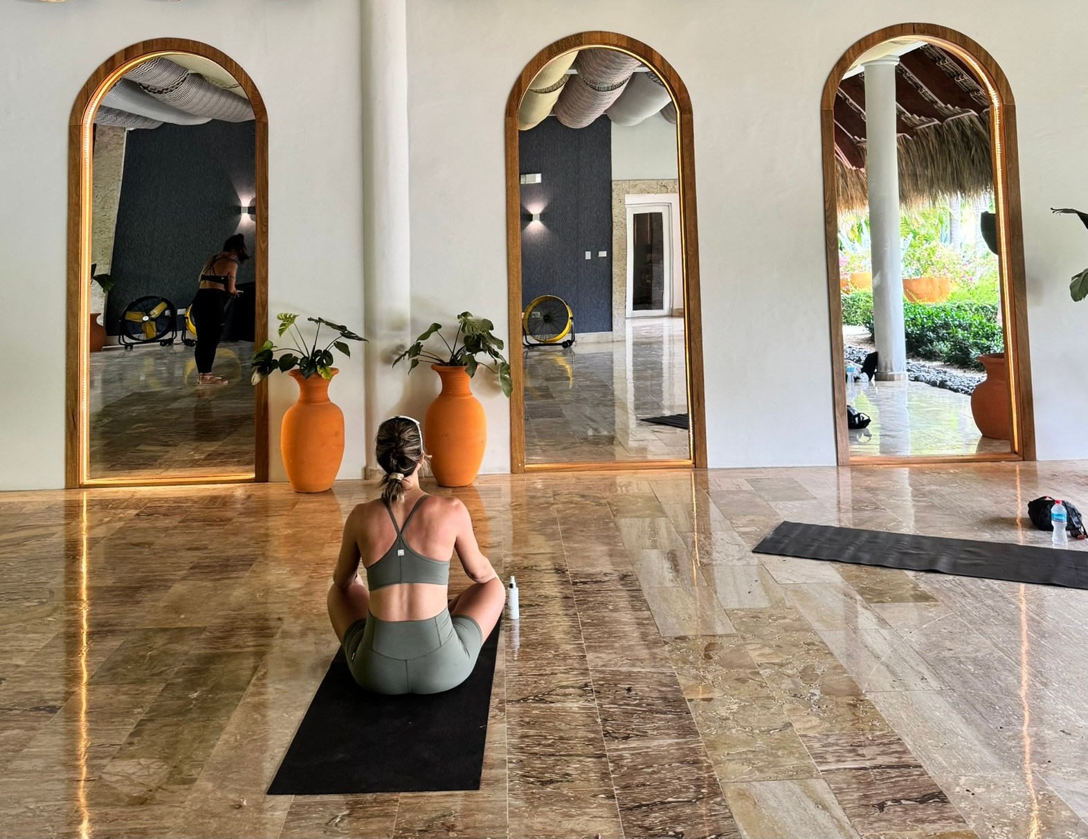 Woman sitting on yoga mat in a studio with patterned fabric rolls on the ceiling and mirrors reflecting the outdoor space.