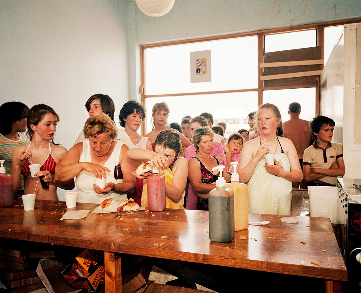 A crowd of people queueing at a counter, adding condiments to their hot dogs and drinks.