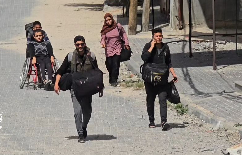 A group of people, including a person in a wheelchair, walk down a street carrying luggage.