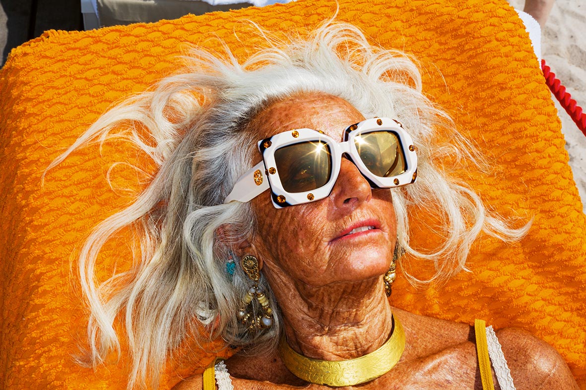 A sun-kissed older woman wearing large sunglasses lies on an orange towel.