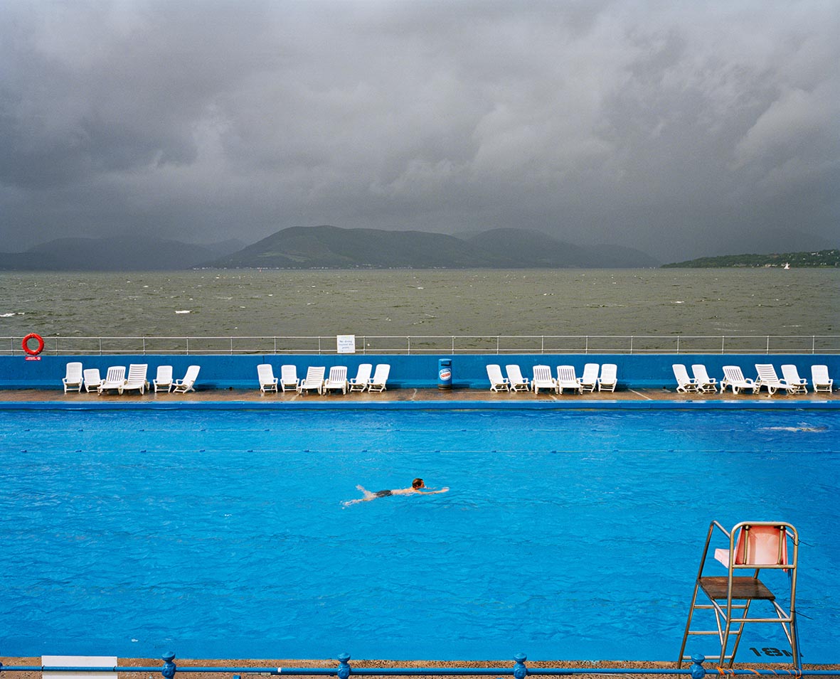 Person swimming in an outdoor pool overlooking a choppy sea and mountains under a stormy sky.