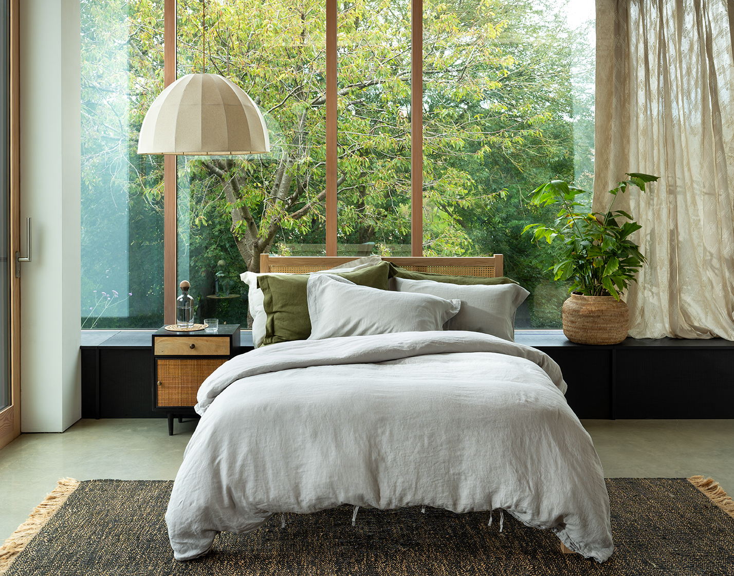Bedroom with light gray bedding, wooden headboard, and large window overlooking a garden.