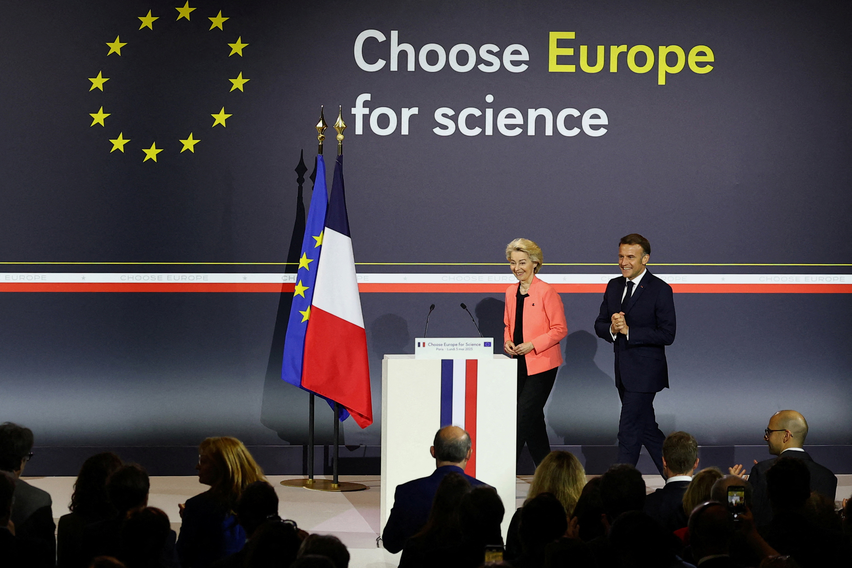 Ursula von der Leyen and Emanuel Macron next to EU and French flags on a stage