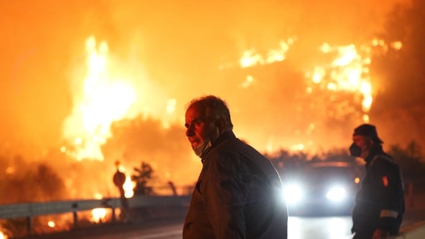 a two men and an approaching car with a railing and wildfires in the background