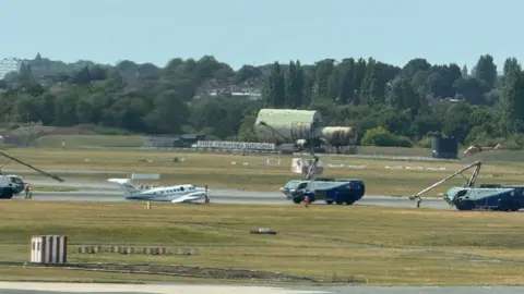 Mr Singh Small white plane on runway surrounded by large vehicles with small crane arms on them.