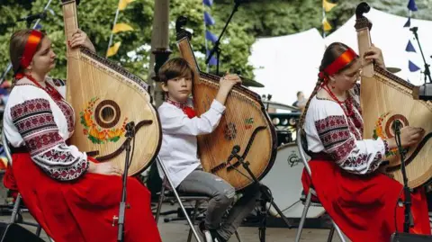 Anastasiia Moskalenko Three musicians at 2024's UkraineFest sat in a row in traditional dress of red and white preparing to play large stringed instrument.