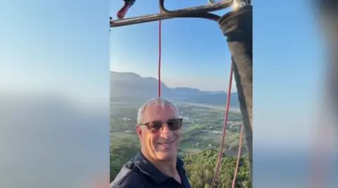 Flying Enterprises Ian Martin pictured in a hot air balloon flying in the sky. He has short grey hair and is wearing dark sunglasses. He is looking at the camera and smiling.