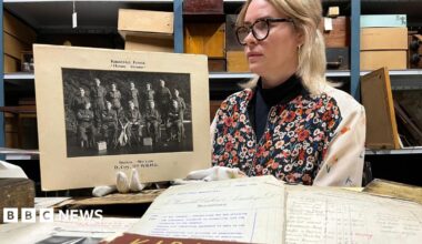 A woman with blonde hair and glasses holds a black and white photograph wearing white gloves. In front of her are a selection of handwritten documents