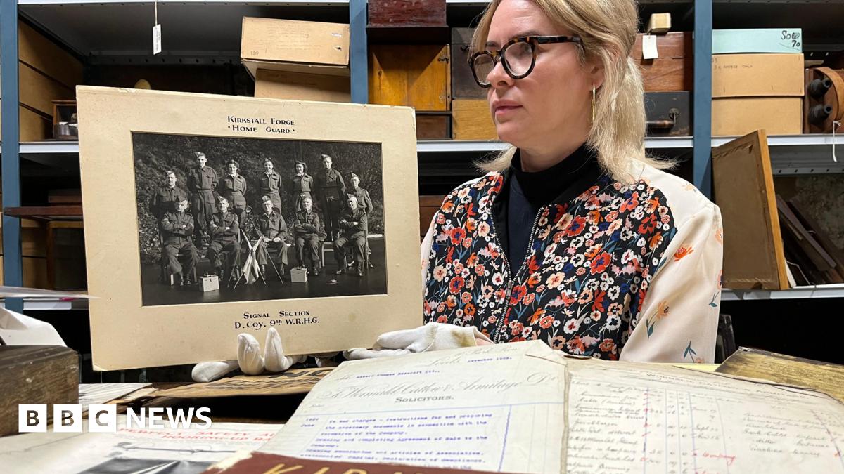 A woman with blonde hair and glasses holds a black and white photograph wearing white gloves. In front of her are a selection of handwritten documents