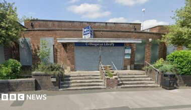 1930s low brick building with steps leading up to it from the road. The entrace is covered in a metal barrier, with a sign above it saying "Pugh Auction". The windows are also covered in grey metal and there are trees and bushes in front of the building.