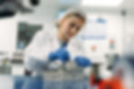 Lab technician in white coat and hairnet, focusing on a metal piece at a blue workbench. Wearing blue gloves in a bright lab setting.