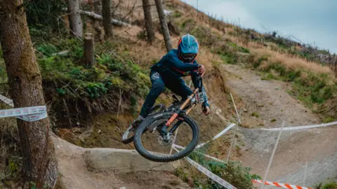 Family photo Ioan Hepburn in his teens competing in a downhill mountain bike race. The location is forestry with a twisty rutted track going between the trees, some of which have been felled. 
He's taking a jump on his bike which is turned almost completely on its side in the air, and is wearing a light blue helmet.  