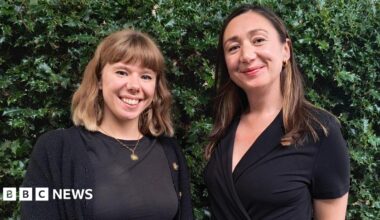 Two women standing in front of a short wall and a holly bush. Both are wearing dark clothing, have brown hair and are smiling.