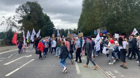 A group of people waving flags leave the side of Ormeau Park, on the Ormeau Embankment.
