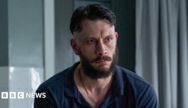 Actor Richard Gadd in a hospital scene in his new series Half Man - he sits looking morose on a dishevelled hospital bed, wearing a blue polo shirt and jeans. He is muscular and has dair hair, a beard and a moustache.