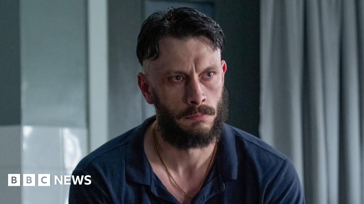 Actor Richard Gadd in a hospital scene in his new series Half Man - he sits looking morose on a dishevelled hospital bed, wearing a blue polo shirt and jeans. He is muscular and has dair hair, a beard and a moustache.
