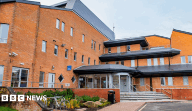 The offices of Tewkesbury Borough Council. It is a large orange brick building with lots of windows and stairs leading up to the coved glass entrance, where there are automatic doors. There is a section of greenery beside a low brick wall, and a small bike rack.
