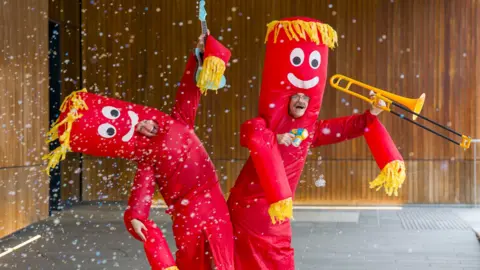 PA Media Two people in red tube costumes with yellow fringing. One holds a violin and the other holds a trombone. Confetti erupts before them and one strikes a jaunty angle.