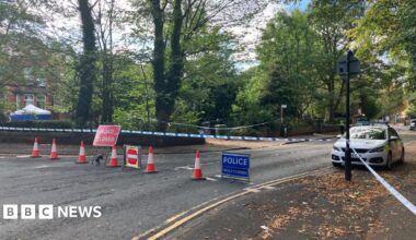 A street is taped off by police, with a blue forensic tent seen in the distance near a house. A police car is parked on the right. The street has trees on both sides, with some leaves on the ground.