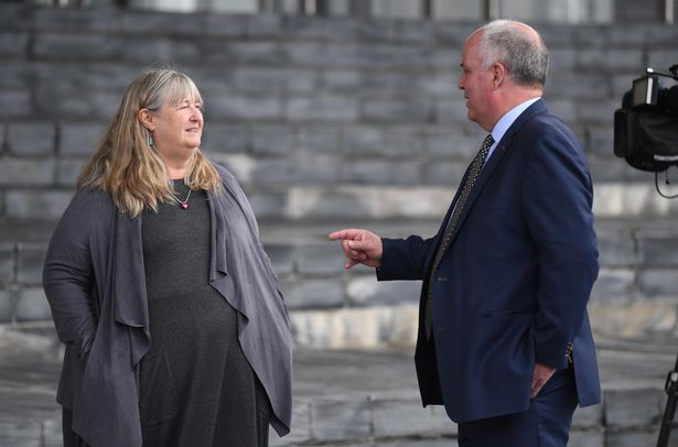 A woman in a black dress and grey cardigan talking to a man in a suit who is pointing