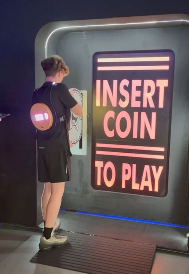 A player is tasked with putting a large fake coin into the 'insert coin to play' slot 