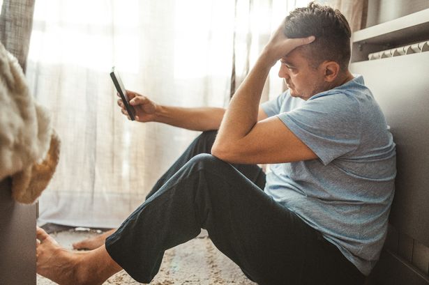 Stressed out middle aged man sitting on the floor of his home, holding a mobile phone