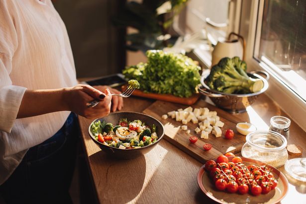 Healthy dinner or lunch. Woman in t-shirt and jeans standing and holding vegan superbowl or Buddha bowl with hummus, vegetable, salad, beans, couscous and avocado and smoothie in hands, square crop