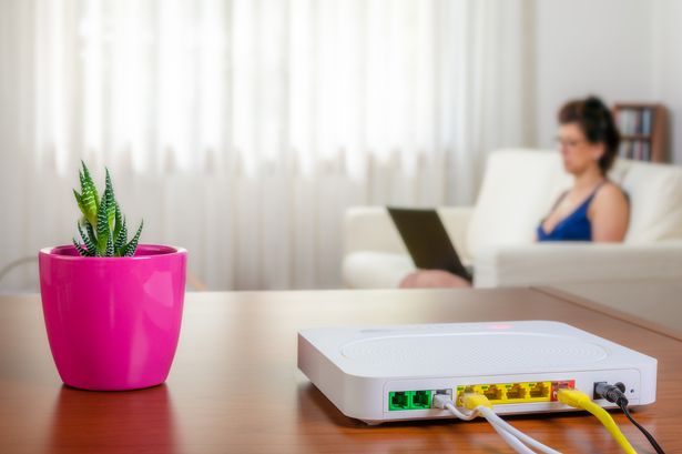 Modem router on a table in a living room. A woman using a laptop while sitting on the sofa is in background.