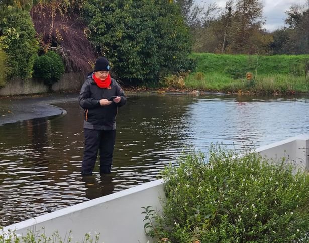 Mournes Alliance councillor Jill Truesdale at a flooded home in Newcastle. She is standing in a body of water and wearing dark water proof clothes abd a red scarf whilst using her phone to text. 