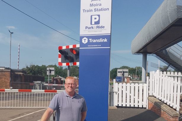 Cll Alan Martin stands with his hand on a railing infront of a vertical sign for Moira train station. 