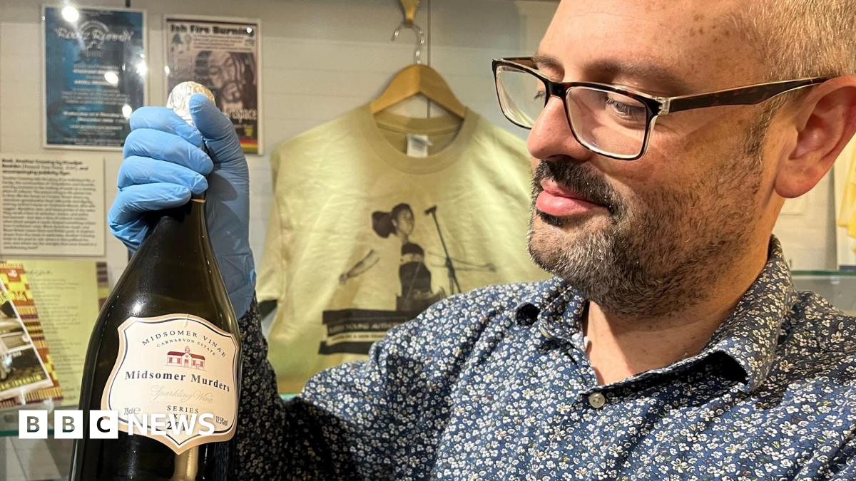 A man with short dark beard wearing a floral blue shirt holds a wine bottle.