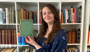 Caitlin Riley is standing in front of a book case holding up the first edition of The Hobbit. It is a green book with a drawing of a dragon on the front. Caitlin is wearing a blue dress and has long, brown hair.