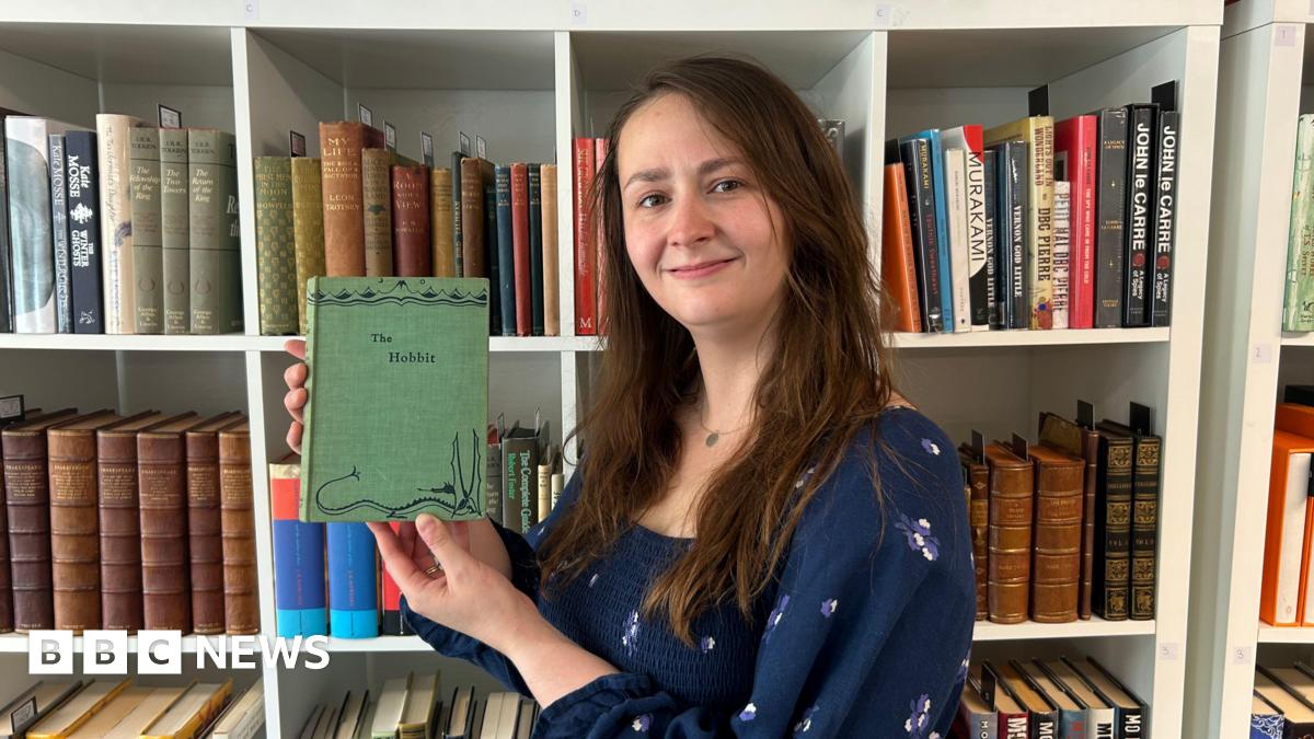 Caitlin Riley is standing in front of a book case holding up the first edition of The Hobbit. It is a green book with a drawing of a dragon on the front. Caitlin is wearing a blue dress and has long, brown hair.