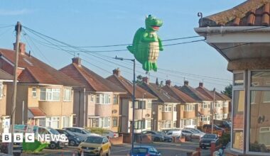 A large dragon-shaped hot air balloon is flying over residential streets with cars lined up.