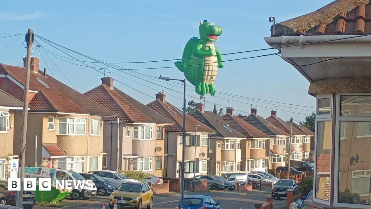 A large dragon-shaped hot air balloon is flying over residential streets with cars lined up.