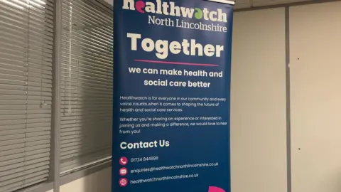 Healthwatch North Lincolnshire sign encouraging the public to get in touch with them so that they can make a difference to health and social care in the local area. It has a blue background and is in an office.