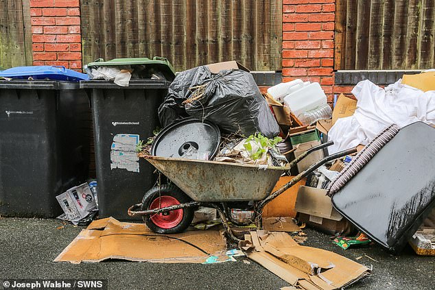 A wheelbarrow takes up space on the pavement besides black wheelie bins and a mound of plastic bags