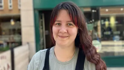 BBC Sian has brown hair with red highlights on her fringe is wearing a grey blue top and black dungarees over it.  