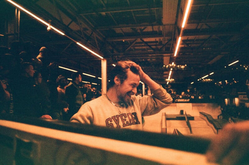 A man in a light sweatshirt smiles and runs his hand through his hair at an indoor skatepark. The background shows spectators and ramps under bright, industrial lighting. The photo has a warm, grainy effect.