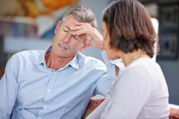 A man and woman are sitting on a sofa, the man has his eyes closed and is holding his hand to his head 