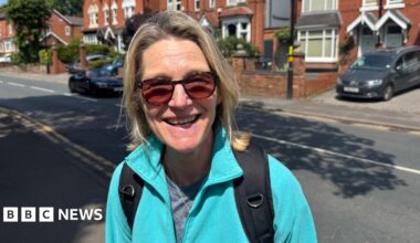 Dee Lyons in a smiling photo. There is a road behind her with a car driving past. She is wearing a light blue fleece and sunglasses. She has blonde hair.