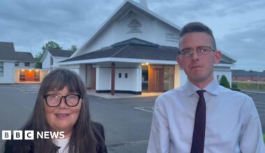 Martina Burke and Josiah Burke standing outside the Hebron Free Presbyterian Church in Ballymoney. Martina has long, dark, greying hair with a fringe, wearing glasses, a black blazer and white top. Josiah has short dark hair, wearing glasses, a white shirt and dark tie. The church is in the background and has a triangular roof.