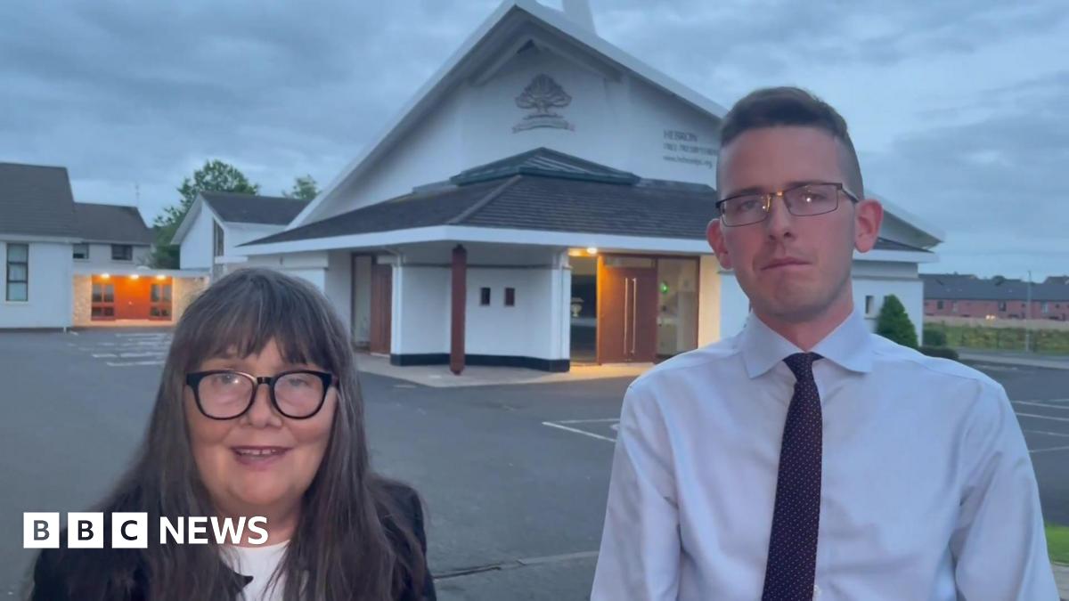 Martina Burke and Josiah Burke standing outside the Hebron Free Presbyterian Church in Ballymoney. Martina has long, dark, greying hair with a fringe, wearing glasses, a black blazer and white top. Josiah has short dark hair, wearing glasses, a white shirt and dark tie. The church is in the background and has a triangular roof.