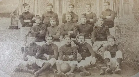Llandovery College A black and white photo of Llandovery rugby team. 
It shows 15 men dressed in long white shorts and long sleeves. 
Some of the men are wearing little floppy hats.