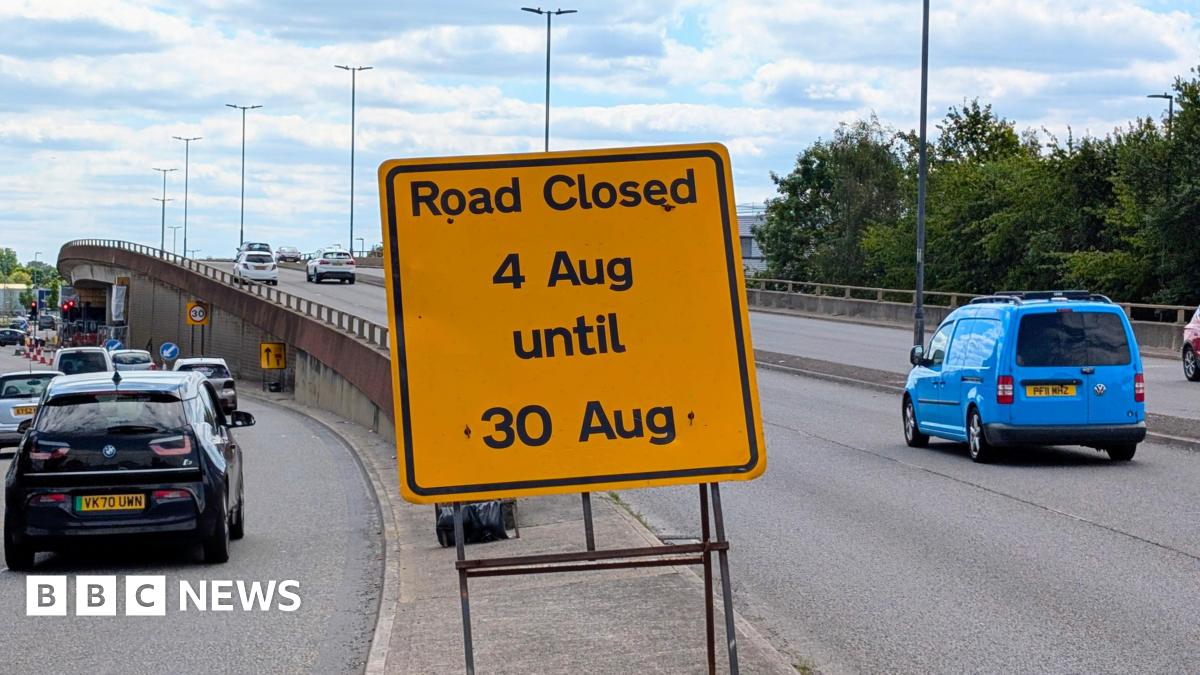 A yellow sign saying road closed 4 Aug until 30 Aug. There is a flyover in the distance with cars driving down the road.