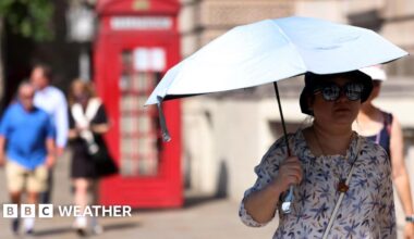 A woman holds an umbrella to shelter from the Sun as she walks down a street. Some people are visible in the background, along with an old-fashioned red telephone box