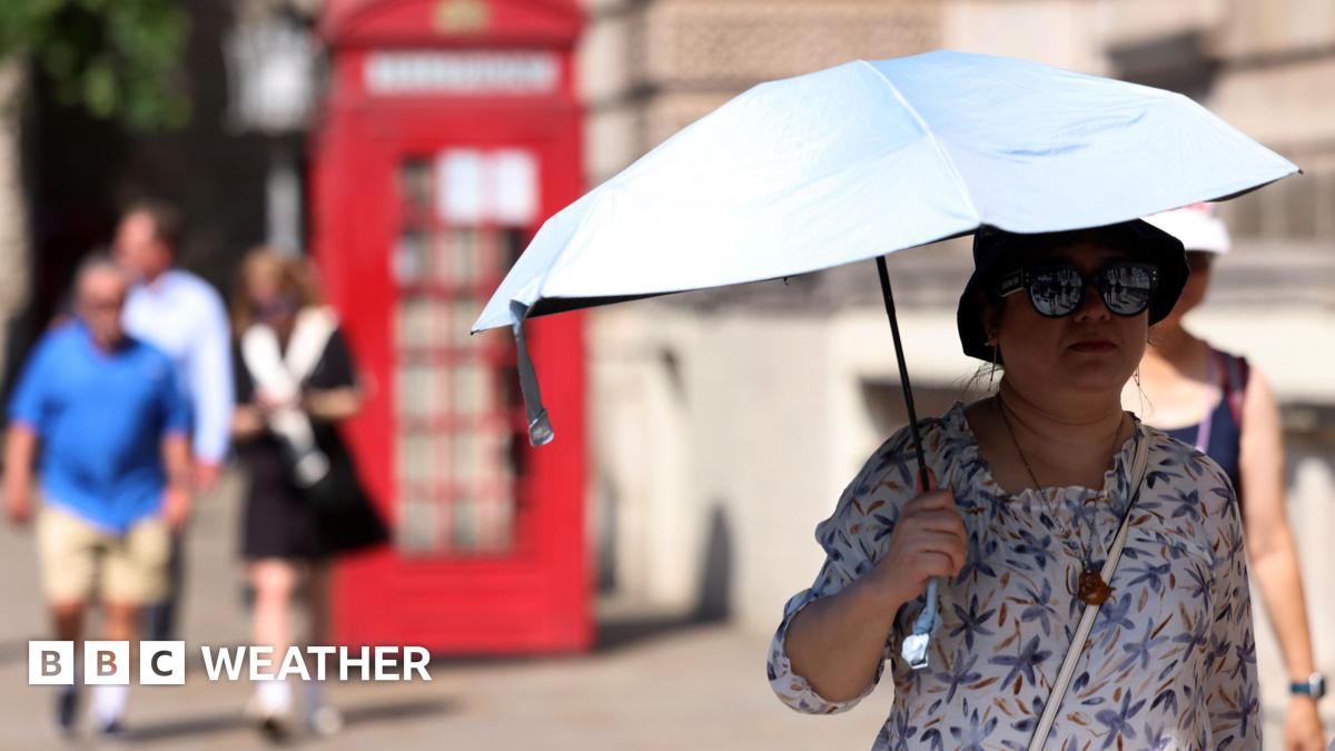 A woman holds an umbrella to shelter from the Sun as she walks down a street. Some people are visible in the background, along with an old-fashioned red telephone box