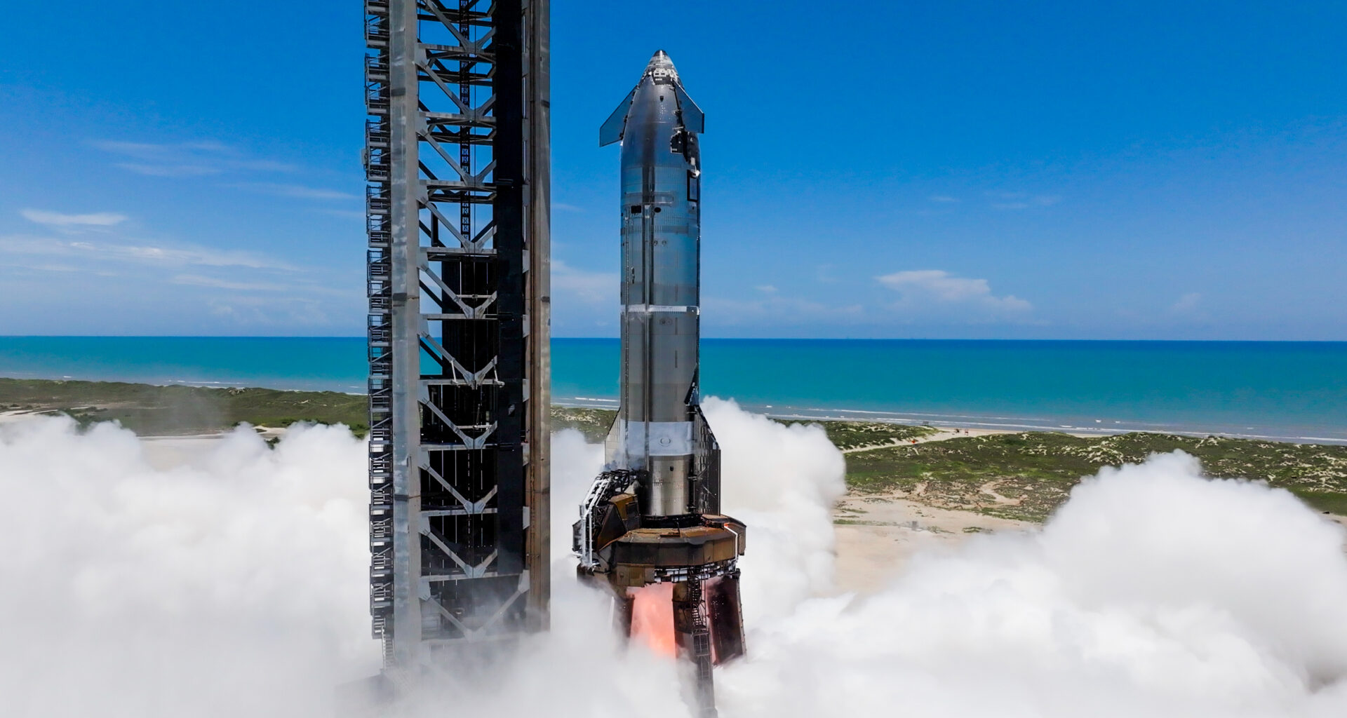 a silver rocket fires one of its engines on a seaside launch pad