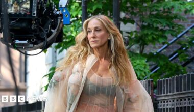 Sarah-Jessica Parker walks along a street with a large camera in front of her.